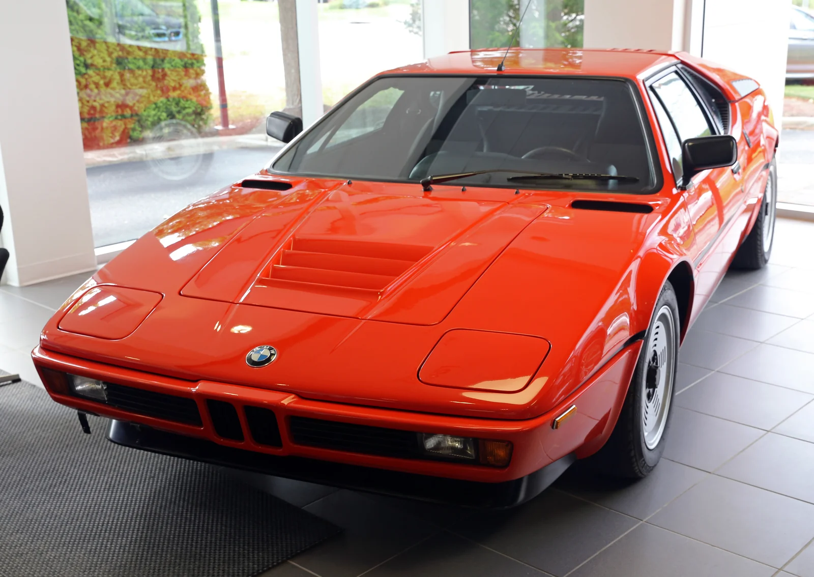 Red BMW M1 E26 on a dealer floor, high front three-quarter showing pop-up headlights and kidney grille
