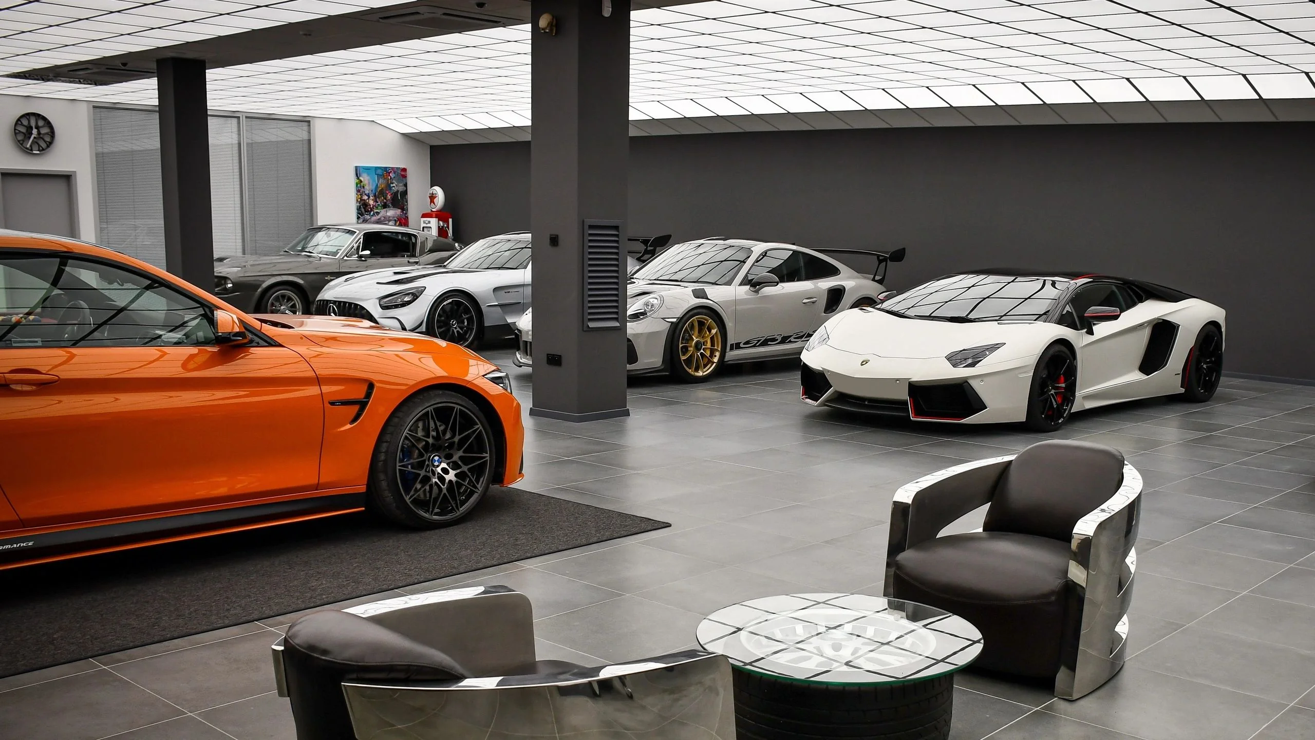 BMW M4 Competition in orange next to Porsche 911 GT3 RS and white Lamborghini Aventador inside Batman Garage