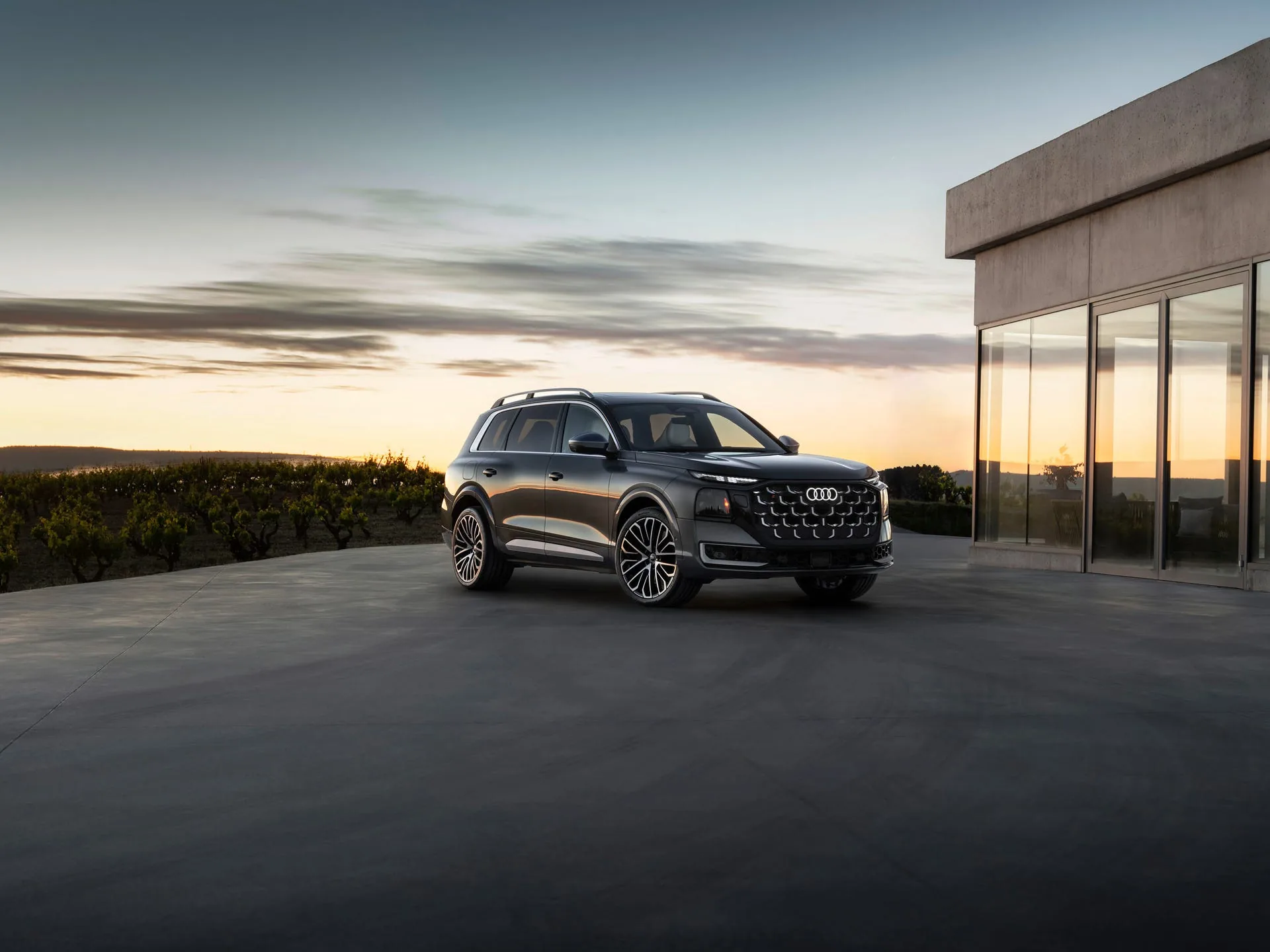 Audi SQ9 front three-quarter at sunset by concrete building — matrix LED headlights, hexagonal honeycomb grille and quad exhaust silhouetted