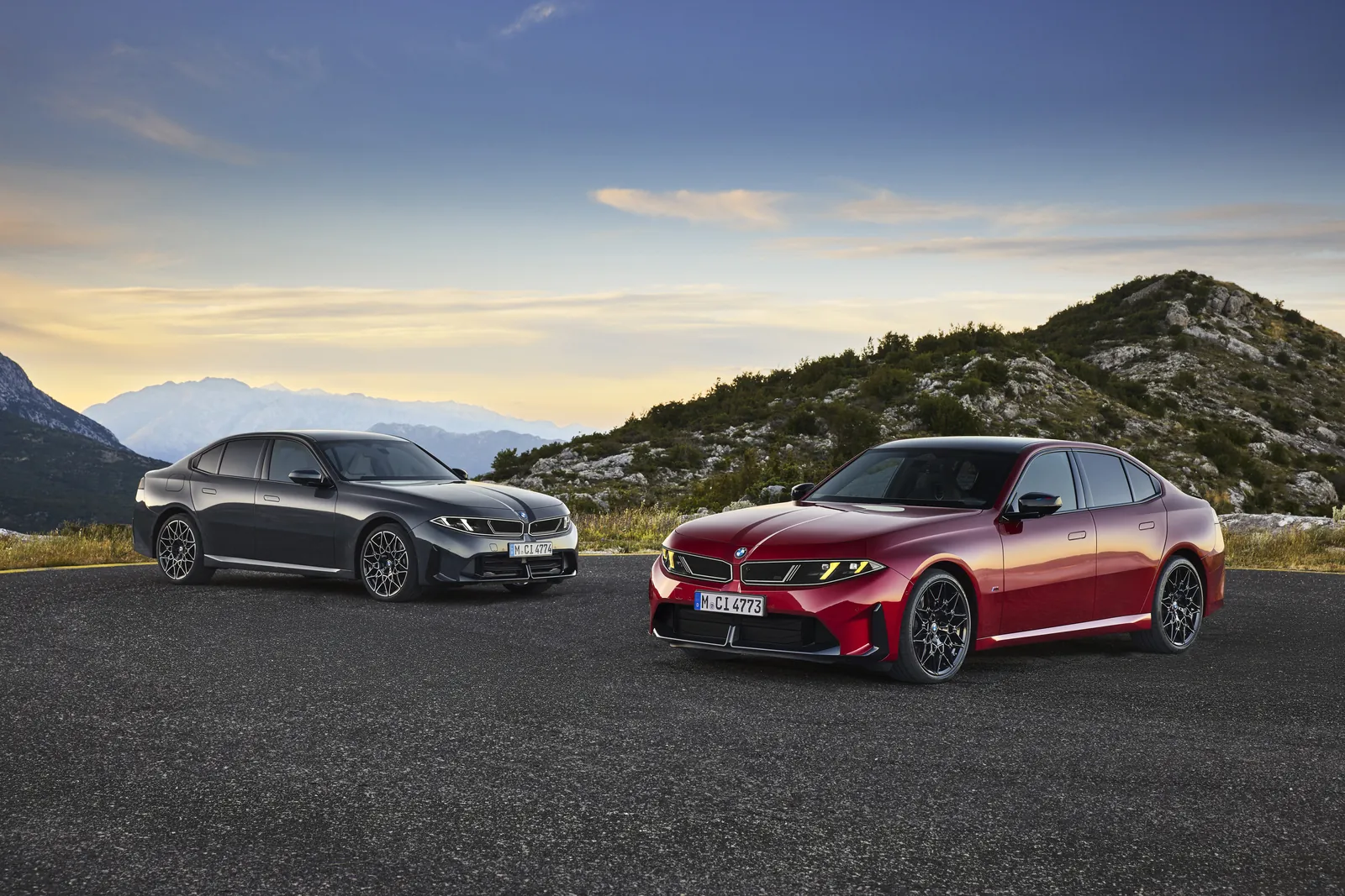 Two new BMW 3 Series G50 sedans in grey and red parked on a mountain road at sunset