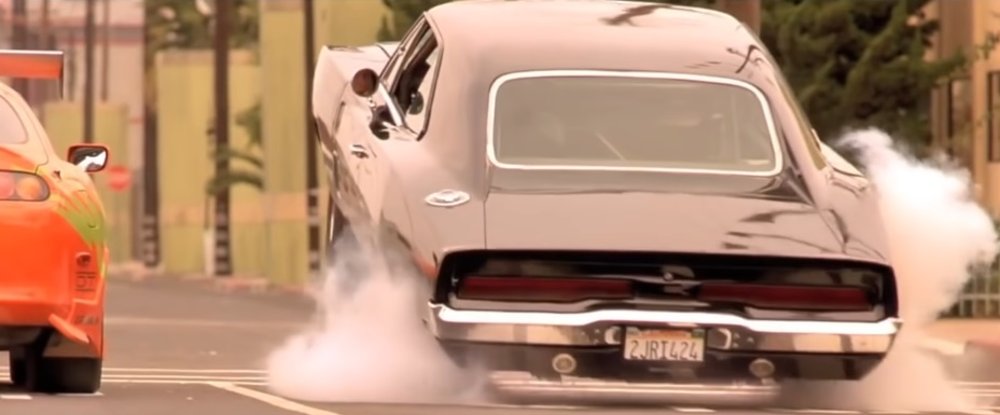 Rear view of Dominic Toretto’s black Dodge Charger smoking its rear tires beside Brian O’Conner’s orange Toyota Supra