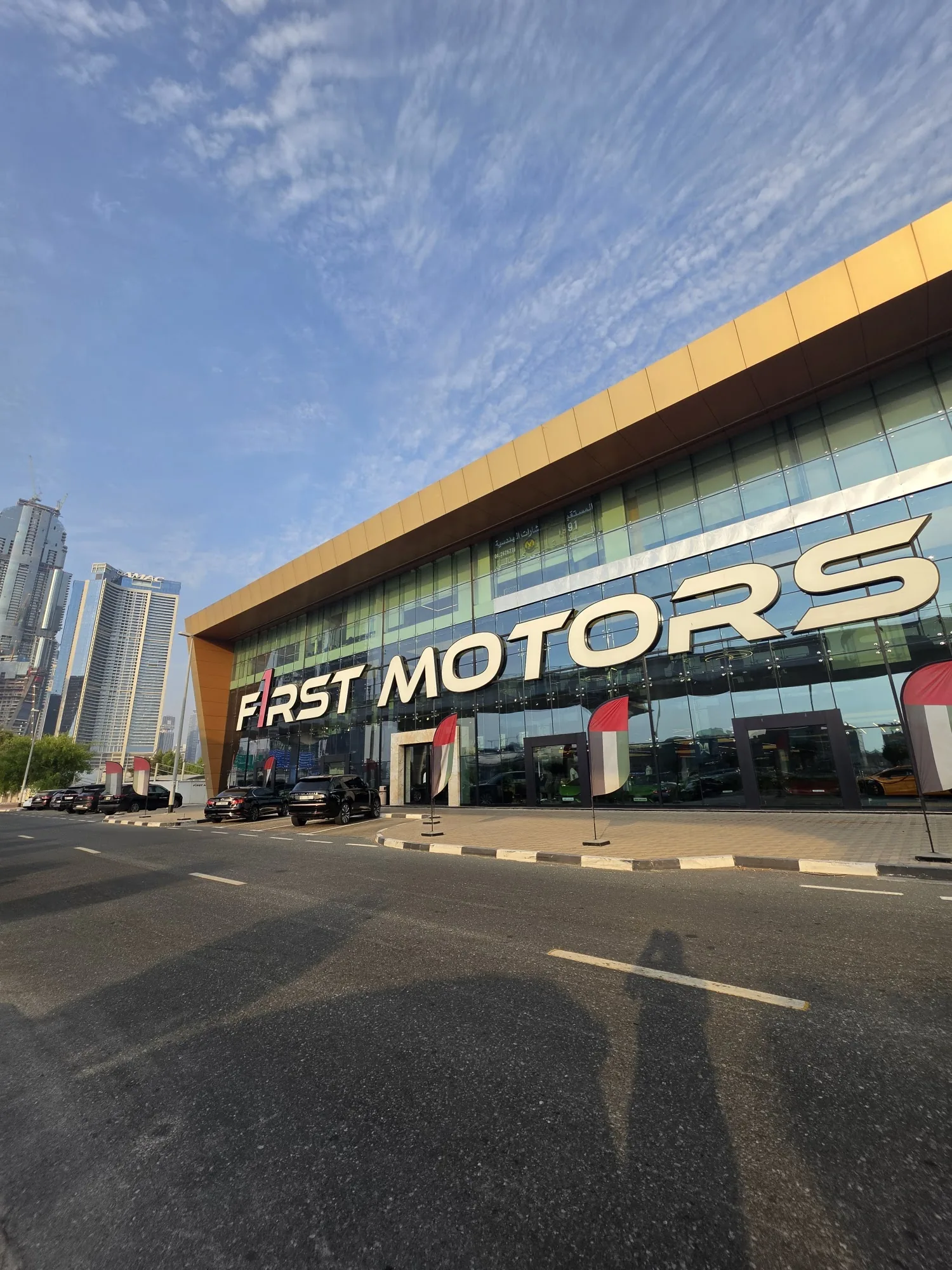 F1rst Motors Dubai exterior facade close-up on Sheikh Zayed Road showing the F1RST MOTORS gold-canopy logo, UAE flags and hypercars parked outside
