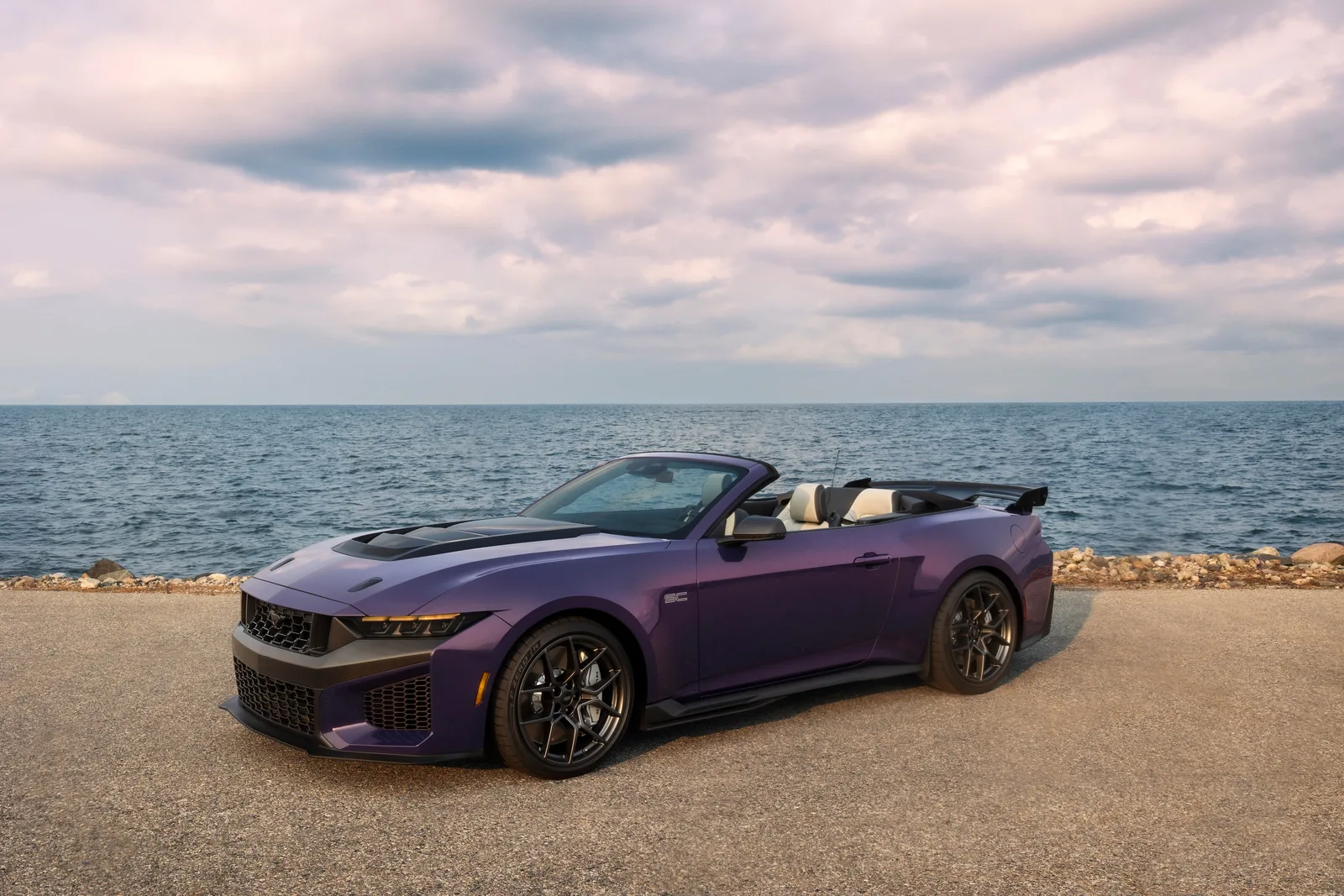 2027 Ford Mustang Dark Horse SC Convertible front three-quarter static view in Precision Purple on lakeside promenade, roof lowered, showing hood scoop, SC-specific front fascia and gloss-black grille