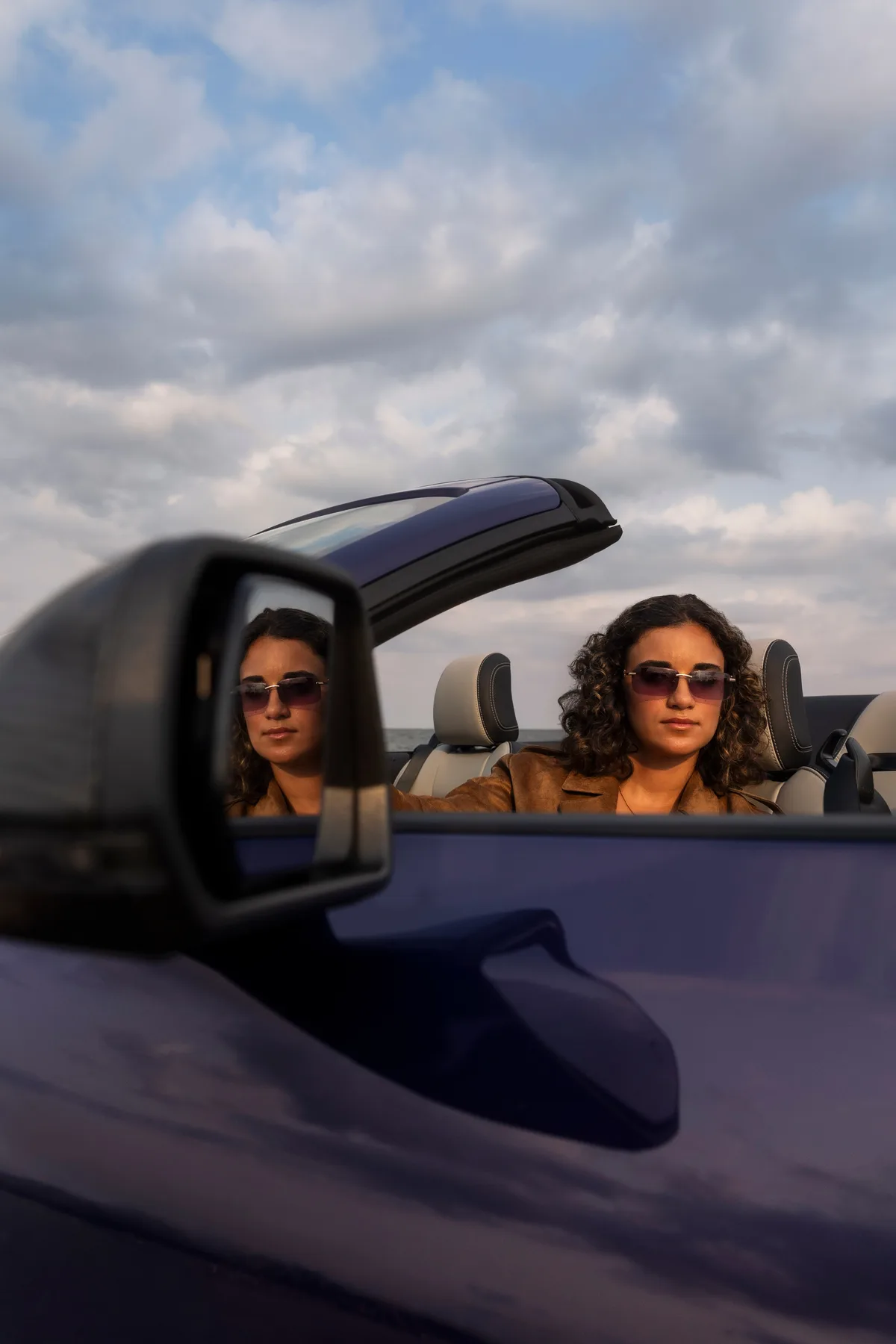Two occupants in the 2027 Ford Mustang Dark Horse SC Convertible with roof lowered, reflected in the side mirror during driving at lakeside sunset