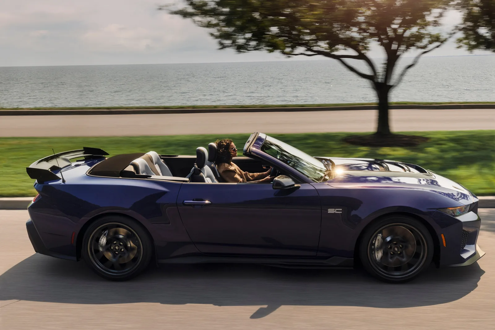 2027 Ford Mustang Dark Horse SC Convertible side profile driving along a lakeside road in Precision Purple, showing full length of the drop-top bodyshell and 19-inch forged wheels