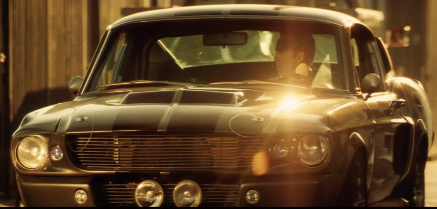 Nicolas Cage driving the Ford Mustang Eleanor at golden hour with lens flare from Gone in 60 Seconds 2000, iconic front three-quarter view of pepper-grey Shelby GT500 tribute