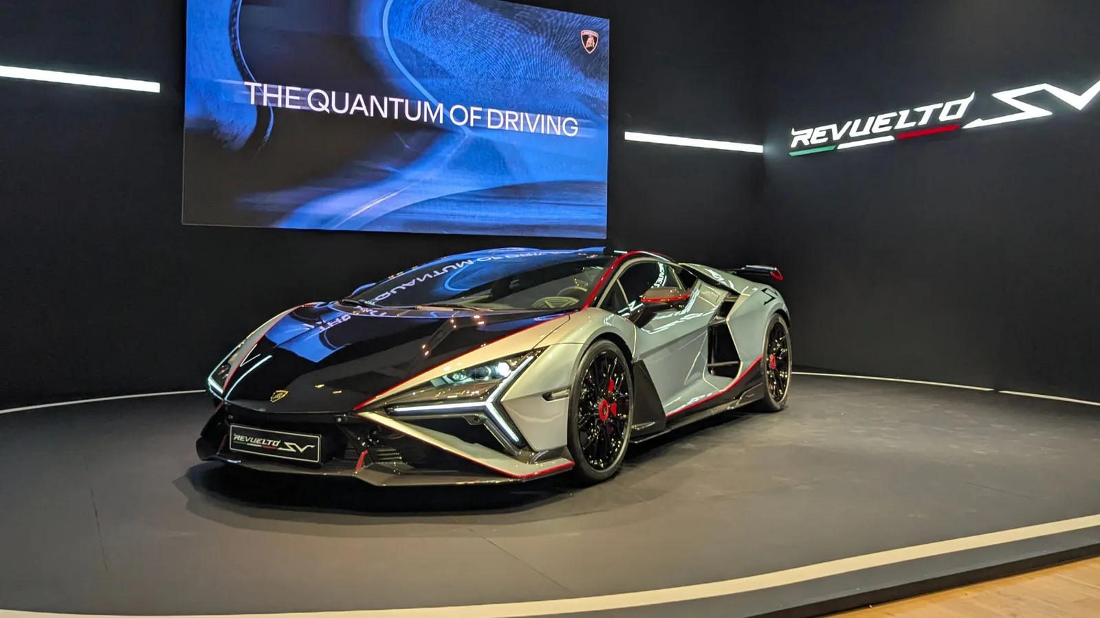 Lamborghini Revuelto SV front three-quarter view at The Quail Monterey Car Week 2026 debut, black-and-silver Rare Silver launch colour with red pinstripe accents, Lamborghini shield illuminated on stand under Quantum of Driving screen