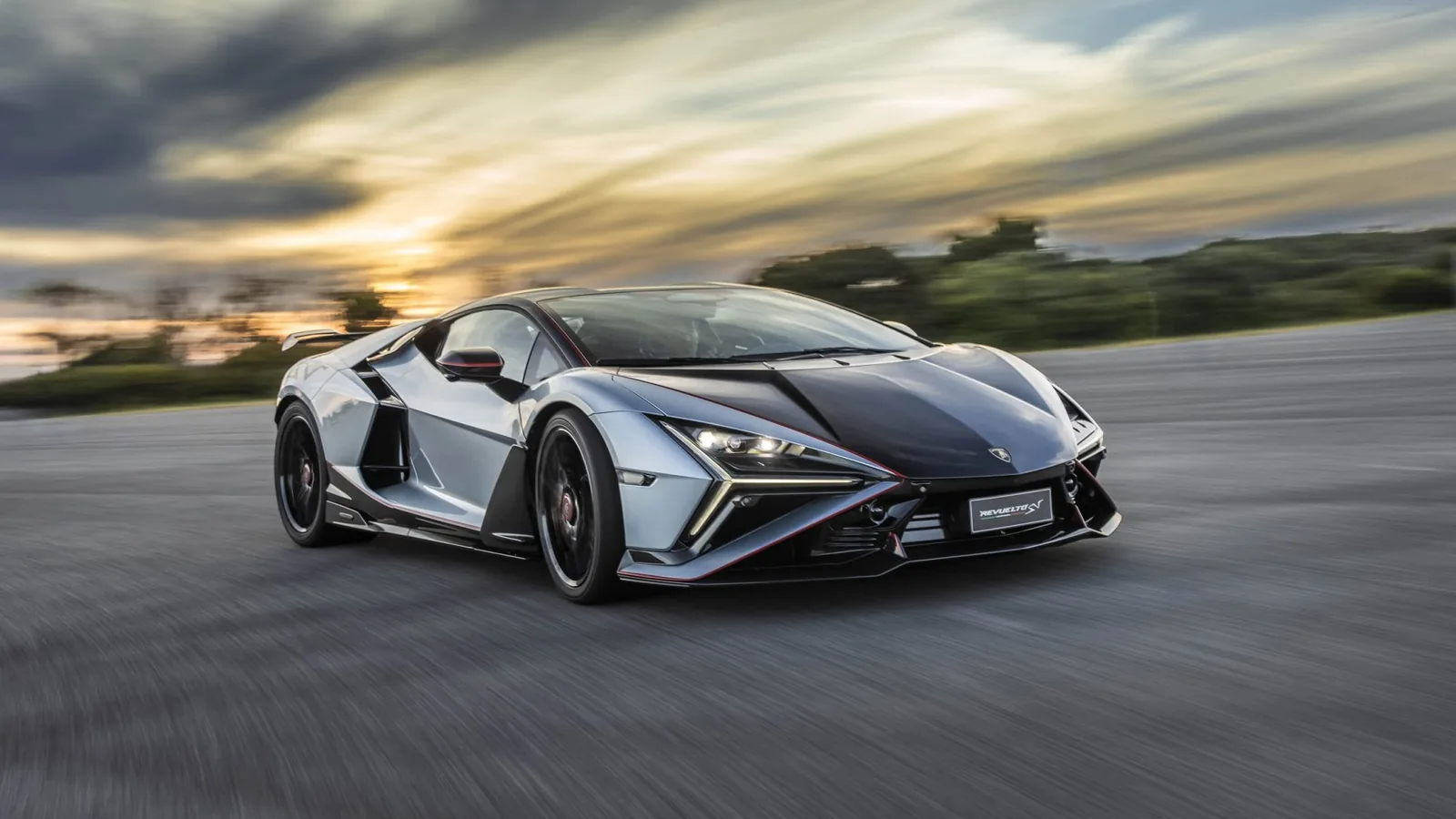 Lamborghini Revuelto SV in motion at dusk showing front three-quarter view, Rare Silver body with black carbon hood and red aero pinstripe, Y-shape LED daytime running lights illuminated