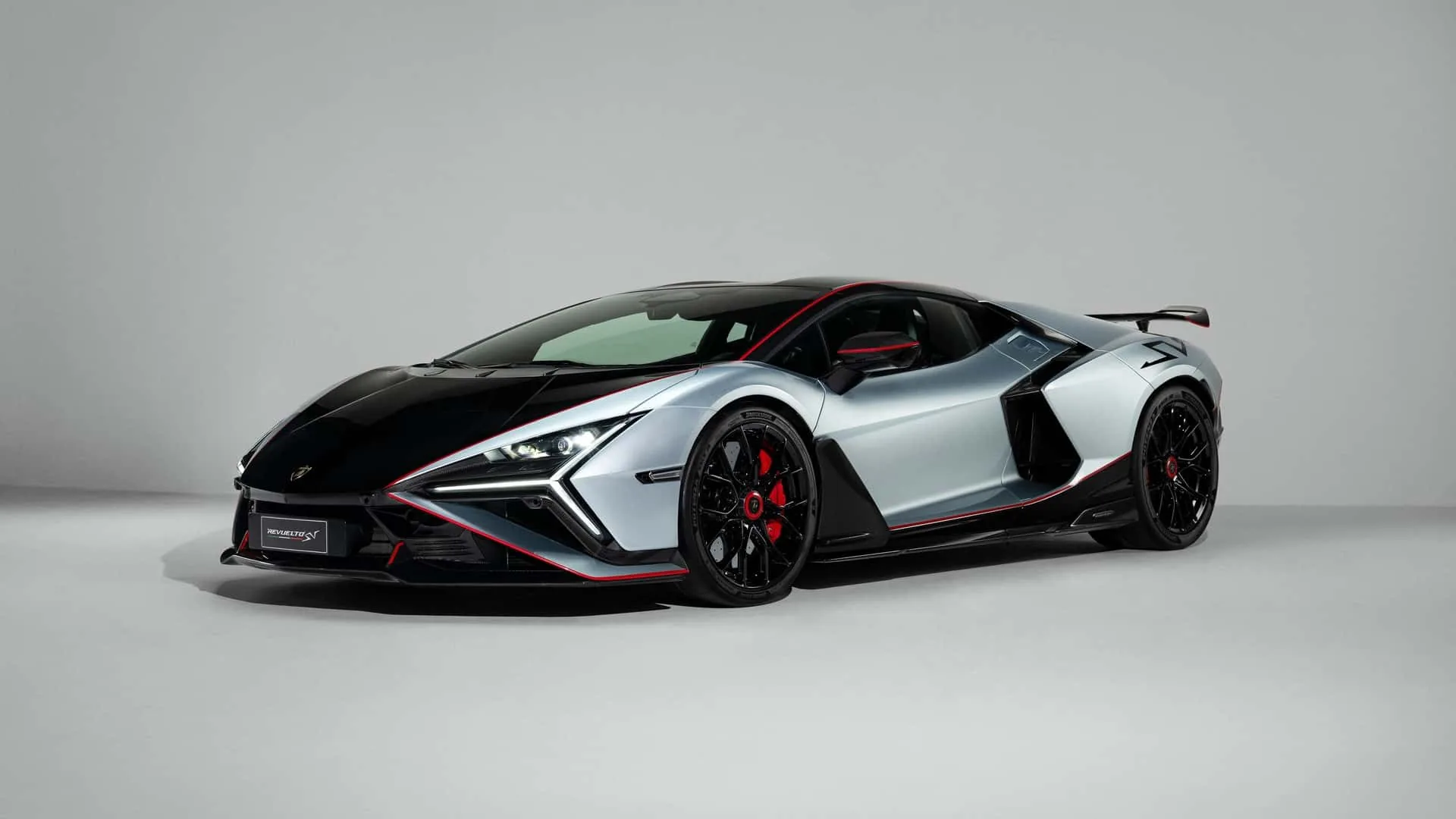 Lamborghini Revuelto SV front three-quarter studio view at Monterey Car Week 2026 reveal, silver-grey V12 hybrid supercar with red pinstripe accents and black hood