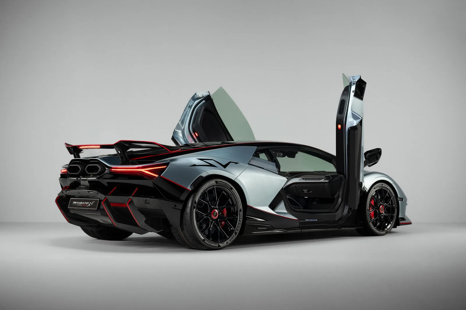 Lamborghini Revuelto SV studio rear three-quarter view with both scissor doors raised, showing full aero package: swan-neck rear wing, quad exhaust, exposed carbon rocker and multi-layer diffuser under Rare Silver bodywork