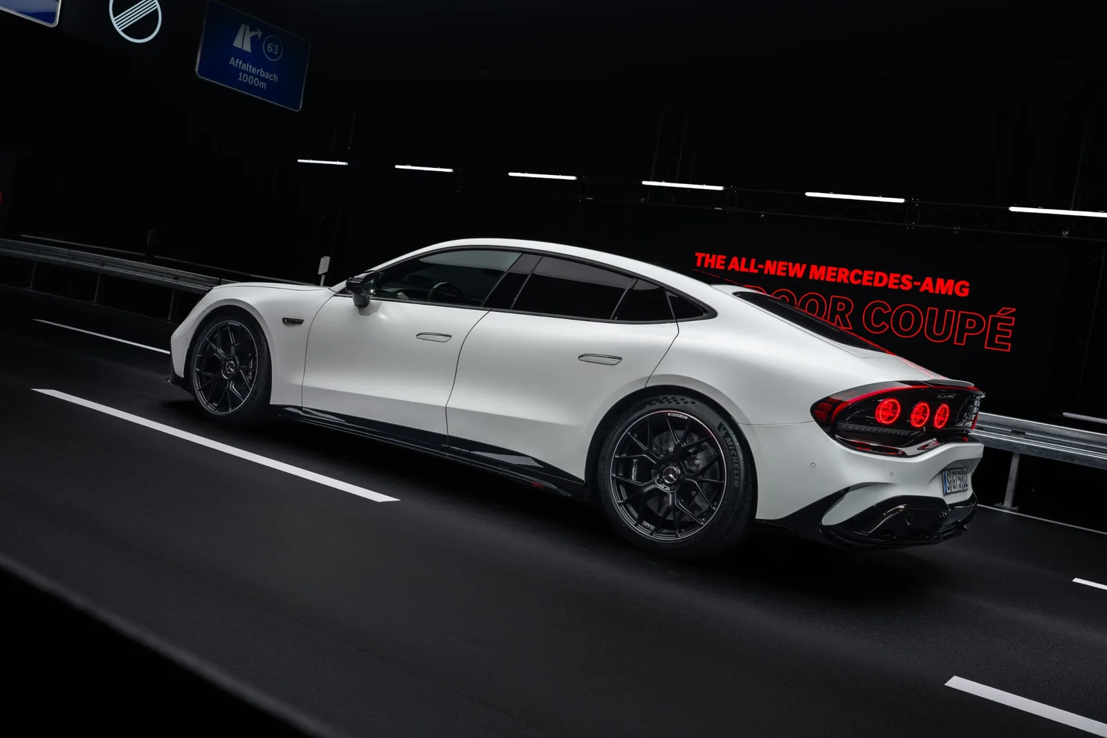 Mercedes-AMG GT 4-Door Coupé EV in white — rear three-quarter side profile against a backlit AMG launch backdrop, showing the 5,094 mm long body, frameless side glass, dark-finish multi-spoke wheels and triple-element circular taillight signature